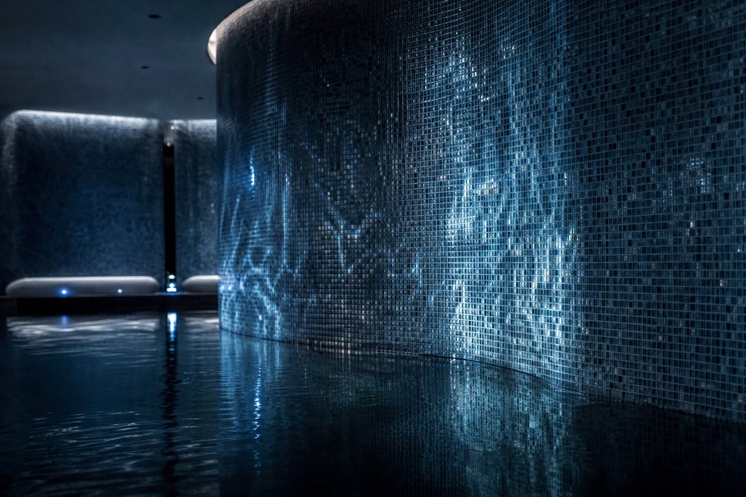 Blue-grey mosaic tesserae on a curved spa pool wall with water reflections.